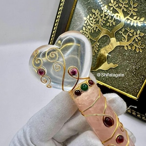 Op de afbeelding: Een hartvormige toverstaf van helder kristal met gouden draaddetails en rode en groene edelstenen. De handgreep is rozenkwarts, omwikkeld met gouddraad. Op de achtergrond een gouden boomontwerp in een lijst.