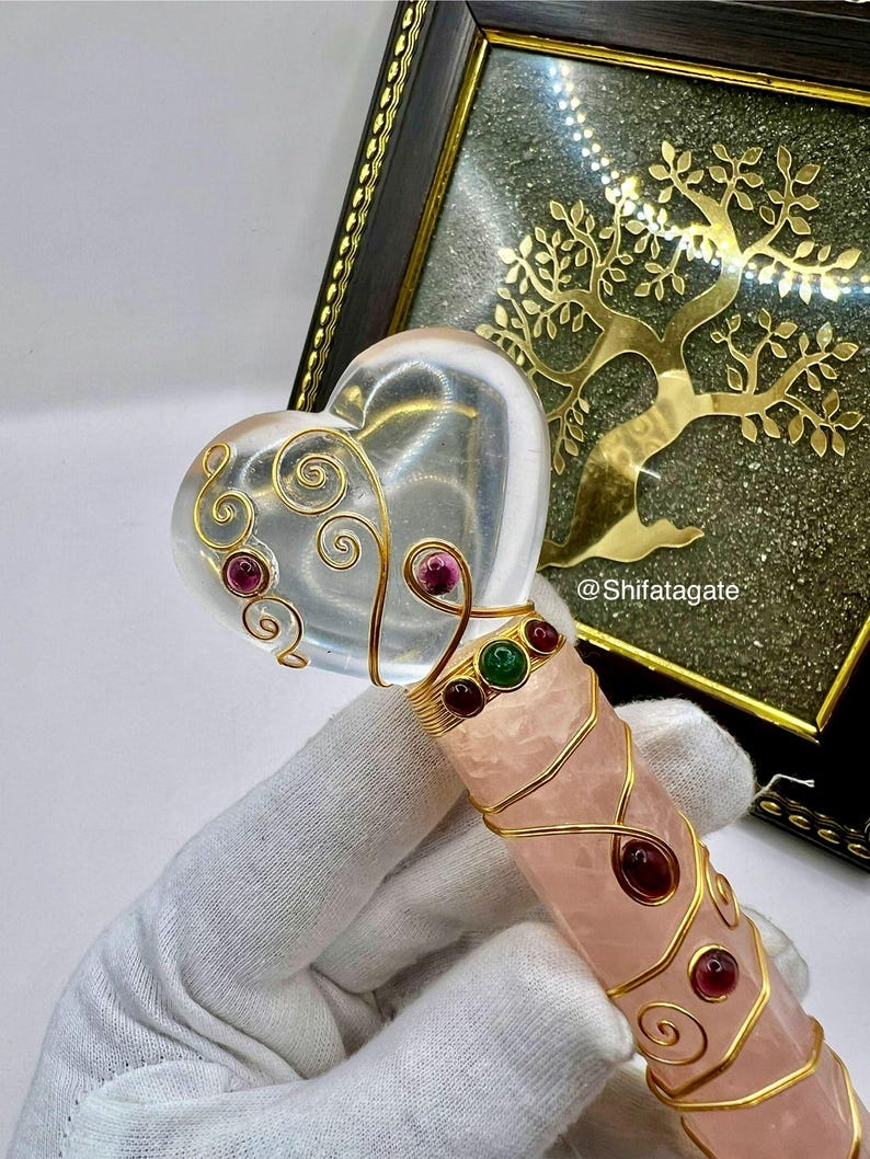 Op de afbeelding: Een hartvormige toverstaf van helder kristal met gouden draaddetails en rode en groene edelstenen. De handgreep is rozenkwarts, omwikkeld met gouddraad. Op de achtergrond een gouden boomontwerp in een lijst.
