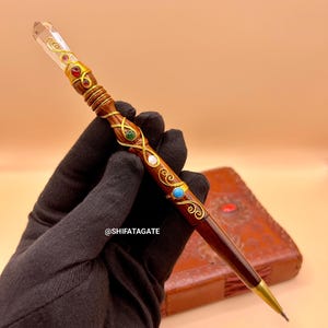 May include: A handcrafted wooden pen with a clear crystal tip and ornate gold and jewel accents. The pen features a dark brown wooden body with gold filigree and colorful gemstones. The pen is held in a black glove.