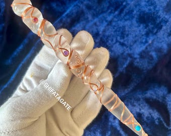 May include: A clear quartz wand wrapped in copper wire with pink, purple, and blue gemstones. The wand is held in a gloved hand. @shifatagate