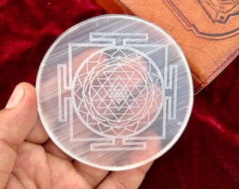May include: A clear, round selenite coaster with an etched Sri Yantra design. The coaster is held in a hand, with a leather-bound book and red velvet fabric in the background.