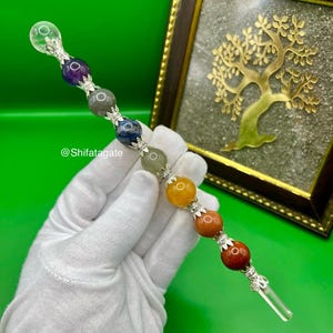 May include: A colorful crystal wand with a clear crystal tip and a series of gemstone beads in various colors, including purple, blue, and orange. The wand is held by a gloved hand. The text "@Shifatagate" is visible.