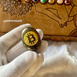 Può includere: Una pietra scura di forma ovale con un simbolo Bitcoin dorato è tenuta in una mano guantata di bianco. Lo sfondo presenta un diario in pelle marrone con dettagli decorativi e il testo "@SHIFATAGATE".