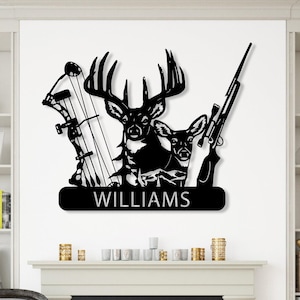 May include: Black metal wall art depicting a deer, rifle, and bow and arrow. The name "WILLIAMS" is displayed on a banner. The artwork is mounted on a white wall above a fireplace in a living room.
