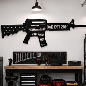 May include: A garage interior with a black metal rifle wall decoration inscribed with "DAD EST 2019". Signs for Ford, Garage, and Texaco are visible. A Chevrolet logo mat is on the floor, and a motorcycle is parked nearby.