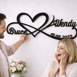 Op de afbeelding: Zwarte metalen wandkunst met een hart en een oneindigheidssymbool. De namen "Bruce" en "Wendy" worden weergegeven, samen met de datum "19.08.2023". Een persoon houdt het kunstwerk vast.