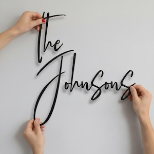 Op de afbeelding: Zwart houten bord in cursiefschrift met de tekst "The Johnsons" tegen een witte muur. Het bord is in een stijlvol scriptlettertype, ideaal voor woondecoratie of een persoonlijk cadeau.
