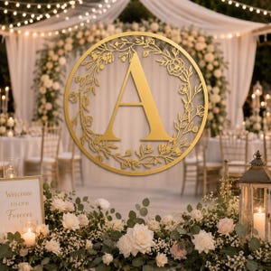 May include: A circular gold-colored wall sign featuring a large capital letter A in the center, surrounded by an intricate laser-cut floral wreath design with delicate stems, leaves, and small blossoms forming a continuous border.