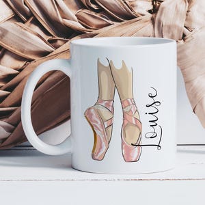 Op de afbeelding: Witte keramische mok met een illustratie van benen en voeten van een balletdanseres in roze spitsschoenen. De naam "Louise" is in cursief schrift geschreven. De mok heeft een wit handvat. De achtergrond is neutraal.