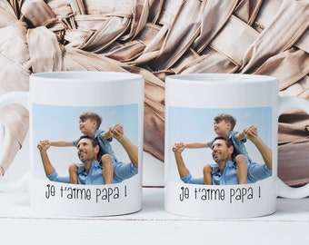 May include: Two white ceramic mugs with a photo of a father and son. The photo shows the son sitting on the father's shoulders. The mugs have the text "Je t'aime Papa!" printed on them.