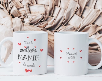 May include: Two white ceramic mugs with black and red text. One mug says "Tu es la meilleure MAMIE du monde" with red hearts. The other mug says "Prenom(s) ou teste" with red hearts. The mugs are on a white surface.