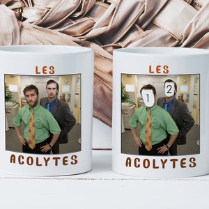 Peut inclure: Deux tasses à café blanches avec une photo de deux hommes dans un bureau. Le texte "LES ACOLYTES" est imprimé sur les tasses. La photo sur l'une des tasses a les visages des hommes remplacés par les chiffres "1" et "2".