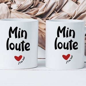 May include: Two white ceramic mugs with black text that reads "Min loute" and a red heart with the text "J'ai kler" below. The mugs are on a white wooden surface.