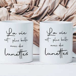 May include: Two white ceramic mugs with black text that reads "La vie est plus belle avec des lunettes" which translates to "Life is more beautiful with glasses".