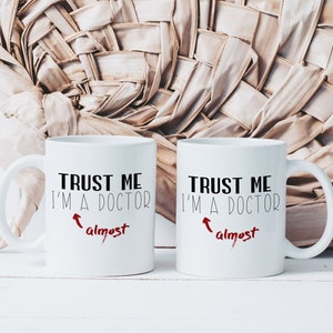 May include: Two white coffee mugs with black text that reads "Trust me I'm a doctor" with an arrow pointing to the word "almost" in red. The mugs are on a white wooden surface.