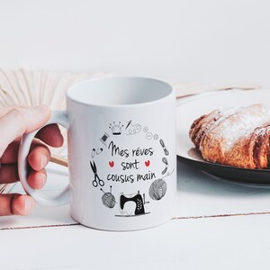 May include: White ceramic mug with a black and red design. The mug has a black handle and the text "Mes r&ecirc;ves sont cousus main" in French. The design includes a sewing machine, yarn, scissors, buttons, and a crown.