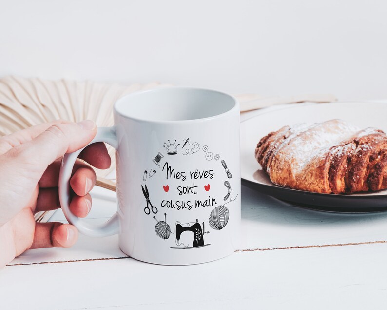 May include: White ceramic mug with a black and red design. The mug has a black handle and the text "Mes r&ecirc;ves sont cousus main" in French. The design includes a sewing machine, yarn, scissors, buttons, and a crown.