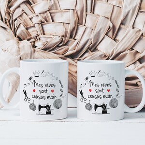May include: Two white ceramic mugs with black text and illustrations. The text on the mugs reads "Mes reves sont cousus main" which translates to "My dreams are hand-sewn". The mugs feature a sewing machine, yarn, scissors, and other sewing-related imagery.