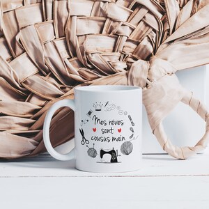May include: White ceramic mug with a black and red design. The mug features a sewing machine, thread, scissors, buttons, and a crown. The text on the mug reads "Mes reves sont cousus main".