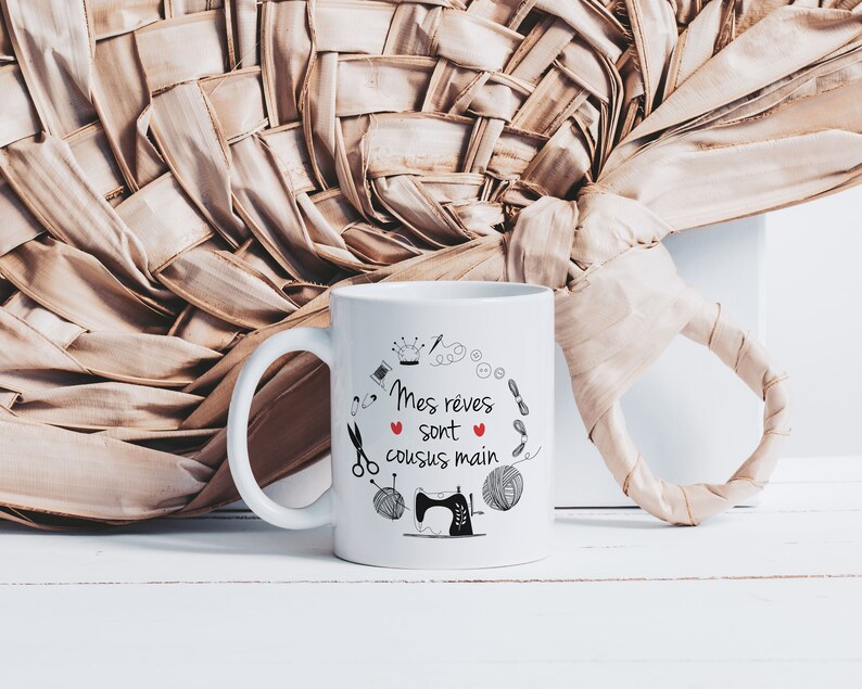 May include: White ceramic mug with a black and red design. The mug features a sewing machine, thread, scissors, buttons, and a crown. The text on the mug reads "Mes reves sont cousus main".