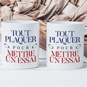 Peut inclure: Deux tasses en céramique blanche avec du texte noir qui dit "Tout Plaquer Pour Mettre Un Essai" avec un symbole de bague rouge de chaque côté du texte. Les tasses sont sur une surface en bois blanc.