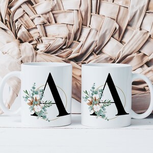 Op de afbeelding: Twee witte keramische mokken met zwarte letter A en gouden cirkel, bloemenontwerp met groene bladeren en witte katoenen bloemen.