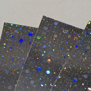 May include: Close-up of several sheets of black, iridescent paper. The paper is covered in a pattern of colorful, shimmering dots and circles in shades of blue, yellow, green, and orange. The sheets are slightly overlapping.