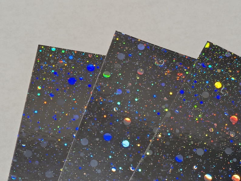 May include: Close-up of several sheets of black, iridescent paper. The paper is covered in a pattern of colorful, shimmering dots and circles in shades of blue, yellow, green, and orange. The sheets are slightly overlapping.