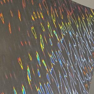 May include: A dark, textured surface with a holographic pattern. The pattern features streaks of color including red, yellow, and blue, creating a shimmering effect. The surface appears to be a large sheet or panel.