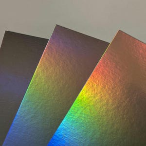 May include: Close-up of three sheets of paper with a holographic finish. The paper edges are visible, displaying a spectrum of colors including blue, green, and orange. The paper has a textured surface.