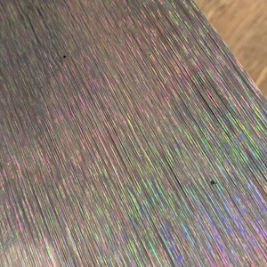 以下が含まれることがあります： ダークグレーのベースカラーを持つ、ブラッシュドテクスチャの金属表面。表面には独特の虹色の効果が現れており、垂直方向の木目全体にピンク、グリーン、ブルーの色合いの繊細で直線的な光の反射が見られます。