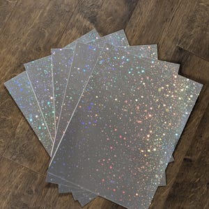 May include: A stack of five rectangular sheets of translucent material featuring a holographic glitter pattern. The surface is covered in small, scattered dots that reflect light in various colors, creating a shimmering, multi-colored effect.