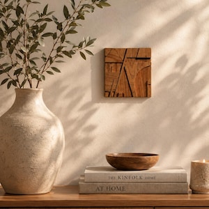 Op de afbeelding: Een vierkant houten wandkunstwerk met geometrisch uitgesneden lijnen en een natuurlijke houtnerf, tentoongesteld naast een grote, getextureerde keramische vaas, een kleine houten kom en twee gestapelde boeken getiteld The Kinfolk Home en At Home.