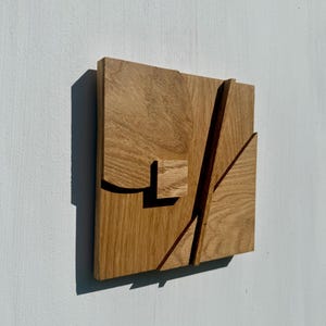 Puede incluir: Una pieza de arte mural cuadrada de madera con veta de roble marrón claro. El diseño consta de formas geométricas superpuestas, incluyendo un cuadrado elevado, una sección curva y listones diagonales que crean profundidad con sombras.