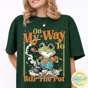Puede incluir: Camiseta verde oscuro con una rana de dibujos animados con peto y una cuchara de madera, con el texto "On My Way To Stir The Pot". La rana está cerca de una olla hirviendo. El diseño es naranja, blanco y verde.