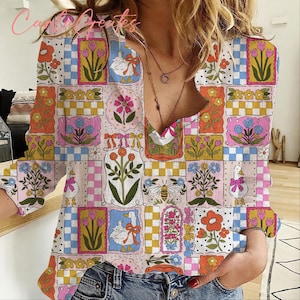 May include: A long-sleeved, V-neck blouse with a patchwork design featuring floral and animal motifs. The pattern includes squares with flowers, ducks, and bees in shades of pink, blue, yellow, and orange. The blouse is worn with blue jeans.