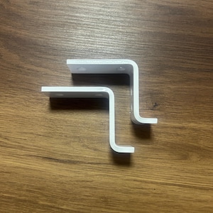 Puede incluir: Dos soportes de estante de metal blanco, cada uno con un ángulo de 90 grados, se muestran sobre una superficie de madera.