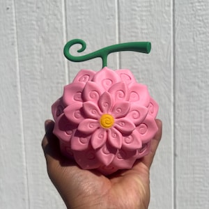 Puede incluir: Un objeto rosa en forma de flor con un tallo verde y un centro amarillo se sostiene en una mano. El objeto tiene un diseño detallado y en capas. El fondo es una superficie de madera blanca.