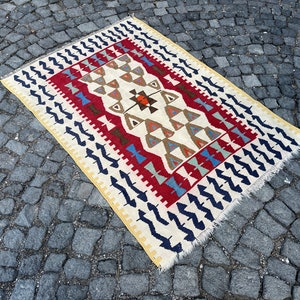 Puede incluir: Una alfombra kilim turca tradicional con un patrón geométrico en tonos de rojo, azul, marrón y blanco. La alfombra tiene un fondo de color crema y un borde con flecos.