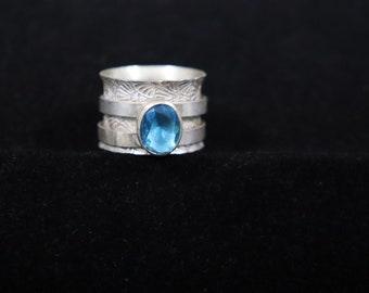 Puede incluir: Un anillo de plata con una piedra preciosa azul engastada en el centro. El anillo tiene una banda texturizada con tres capas.