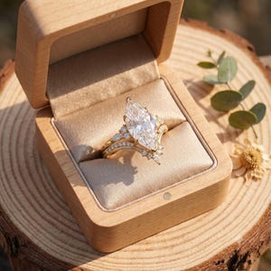 May include: A gold ring featuring a large marquise-cut center stone and smaller accent stones along the band, presented inside a light-colored wooden box with a beige satin interior lining, showcasing a detailed metal setting and clear gemstones.