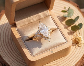 May include: A gold ring featuring a large marquise-cut center stone and smaller accent stones along the band, presented inside a light-colored wooden box with a beige satin interior lining, showcasing a detailed metal setting and clear gemstones.
