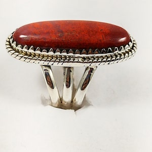 Peut inclure: Une bague en argent avec une pierre ovale rouge sertie dans une bélière en argent. La bague a une bande tressée avec un bord décoratif.