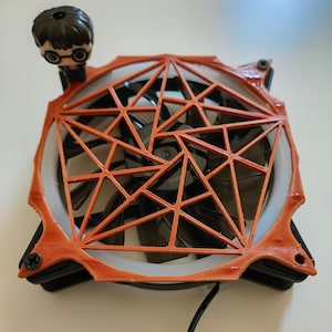 Könnte beinhalten: Ein Computer-Lüfter mit schwarzem Rahmen und dunkel durchscheinenden Flügeln, ergänzt durch ein kupferfarbenes, 3D-gedrucktes geometrisches Gitter. Eine kleine Figur mit dunklem Haar und runder Brille ist an der oberen Ecke befestigt.