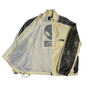 May include: Cream-coloured zip-up jacket with black mesh accents on the sleeves and shoulders. The jacket features an alien graphic and the word "BESCHWA" printed on the front and sleeve.