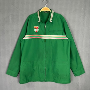 Puede incluir: Chaqueta de trabajo verde con cierre de cremallera y dos bolsillos. La chaqueta tiene una franja blanca, roja y naranja a través del pecho con el logotipo de 7-Eleven.