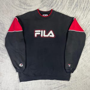 May include: Black FILA sweatshirt with red and white accents. The sweatshirt features a red collar and shoulder panels, with the FILA logo in white with a red outline across the chest. A small FILA logo is on the left sleeve.