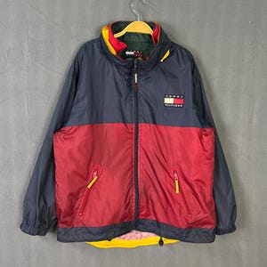 May include: A navy blue, red, and yellow Tommy Hilfiger jacket. The jacket has a full zipper, two front pockets with yellow zippers, and the Tommy Hilfiger logo on the chest. The jacket is hanging on a wooden hanger.