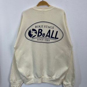 May include: Cream-coloured sweatshirt with a navy blue oval graphic on the back. The graphic reads "BIKE STAGE Be ALL SINCE 1989". The sweatshirt is hanging on a wooden hanger against a plain white wall.