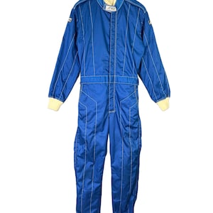 May include: A blue racing suit with white stitching and cream-colored cuffs and collar. The suit has long sleeves and legs, with a zipper closure down the front. The suit has the text "ARD" on the collar.